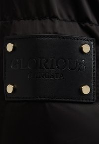 Schwarzes Stoffetikett mit geprägtem Text "GLORIOUS" und "GANGSTA", gesichert mit vier goldfarbenen Metallnieten. Glatte Textur.
