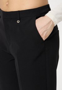 Pantalon noir ajusté avec une texture lisse, doté de poches latérales et d'une accentuation en bouton métallique. Des lignes épurées mettent en valeur un design moderne.