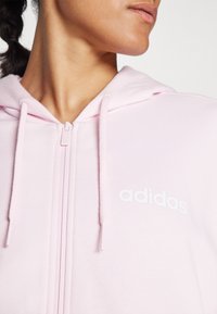 Hele roosa lukuga kapuutsiga sviiter, mis on valmistatud pehmest kangast, mille rinnal on valge "adidas" logo ja kapuutsi juures nöörid.