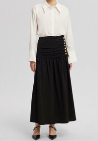 Weißes Hemd mit Kragen kombiniert mit einem schwarzen, gerafften Maxirock mit seitlichen Knöpfen. Das Outfit hat eine glatte Textur und eine strukturierte Silhouette.