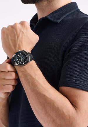 Man adjusting black chronograph wristwatch with silver bezel while wearing a navy blue textured polo shirt.