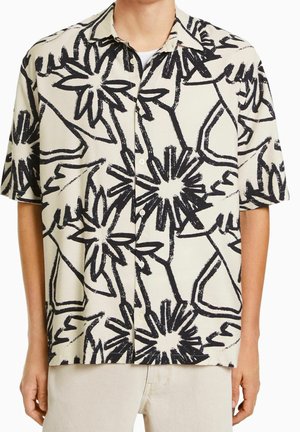 Camicia a maniche corte con bottoni, decorata con motivo floreale astratto nero su sfondo bianco sporco, indossata da una persona con pantaloni beige chiaro.