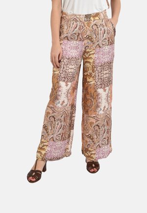 Donna che indossa pantaloni a gamba larga con motivi patchwork paisley multicolori e tacchi aperti marroni, in piedi con le mani nelle tasche.