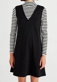 Robe noire sans manches avec un décolleté en V portée sur un col roulé ajusté rayé noir et blanc. Tissu lisse avec une coupe décontractée et un ourlet évasé.