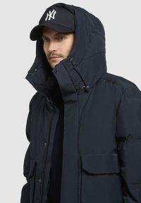 Marine puffer jakke med en stor, justerbar hætte, frontlynlås og sidelommer. Båret med en sort baseballcap med et logo.