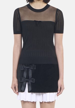 Femme portant un haut en maille noire avec un empiècement et des manches transparentes, assorti à une jupe noire ornée de deux nœuds en satin et d'un ourlet plissé blanc.