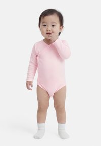 Tout-petit debout, portant un body rose à manches longues et des chaussettes blanches, une main près de la bouche sur un fond blanc uni.