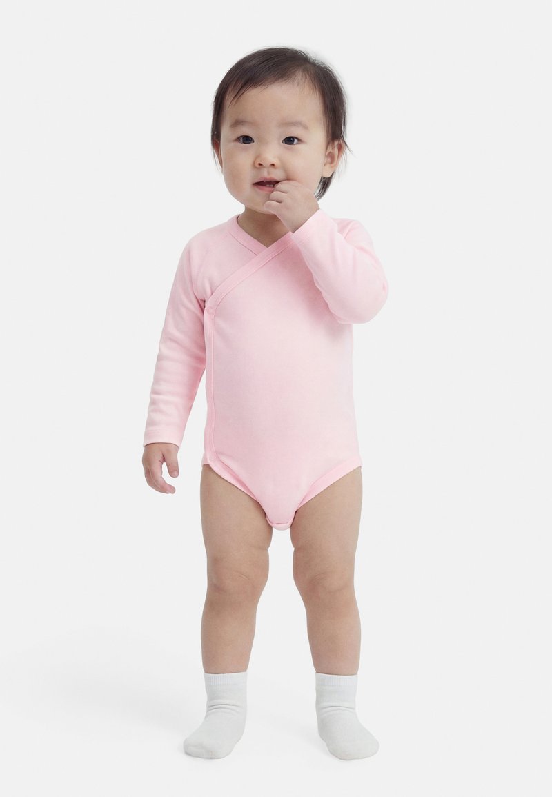 Tout-petit debout, portant un body rose à manches longues et des chaussettes blanches, une main près de la bouche sur un fond blanc uni.