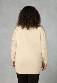 Person with curly hair wearing a beige long sweater with sequin embellishments on the sleeves and black pants, facing away on a gray background.