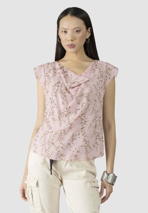 Donna con capelli lunghi e scuri, che indossa orecchini a cerchio, una blusa senza maniche a fiori rosa, pantaloni cargo color crema e un grande braccialetto d'argento.