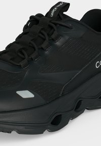 Μαύρο αθλητικό παπούτσι Calvin Klein με χοντρή σόλα, δεμένα κορδόνια, ανακλαστική λεπτομέρεια κοντά στα δάχτυλα και το λογότυπο Calvin Klein στην πίσω τραβηχτή ταινία και στην πλευρά.