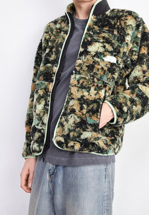 Persona vistiendo una chaqueta de forro polar con estampado en verde, negro y beige sobre una camiseta gris oscuro, con jeans azul claro y la mano en el bolsillo de la chaqueta.