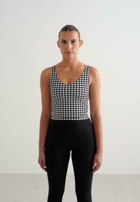 Juodai balta houndstooth raštuota liemenė su plačiomis petnešomis, derinama su siaurais juodais leggingais, turinčiais lygią juosmenį.