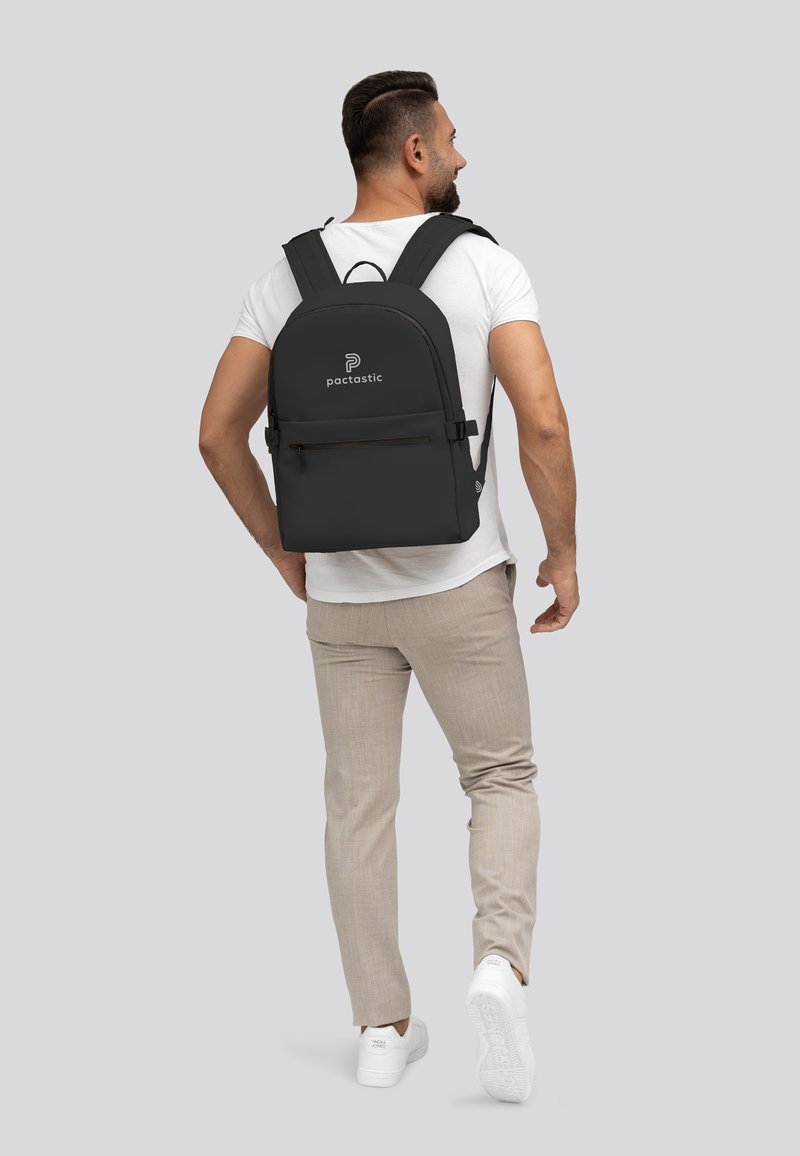 Schwarzer Rucksack mit gepolsterten Trägern, vorderer Reißverschlusstasche und dezentem Logo. Glattes Material und abgerundete Form. Getragen von einem Mann in heller Kleidung.