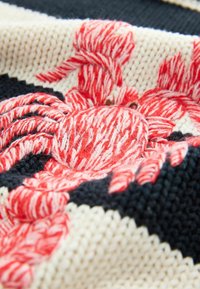 Crabe brodé en fils rouge et blanc sur un fond en tissu tricoté noir et crème.
