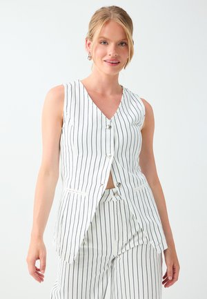 Young woman wearing a white sleeveless V-neck buttoned vest and matching pinstripe wide-leg trousers smiling against a plain background.