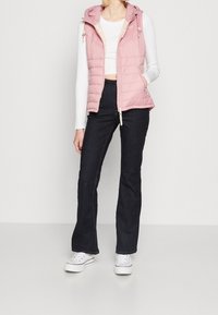 Ljusrosa pufferväst med huva, framdragkedja och sidofickor. Bärs över en vit långärmad topp, i kombination med mörka jeans och vita sneakers.