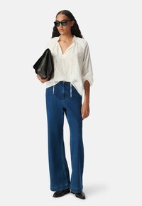 Blusa bianca a pois con una vestibilità rilassata, abbinata a jeans a gamba larga blu. Gli occhiali da sole neri e una borsa a pochette nera completano il look.