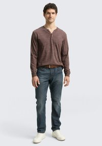 Bordeaux lange mouwen henley shirt met een twee-knopen placket, gecombineerd met blauwe jeans en witte sneakers. Model staat tegen een effen achtergrond.