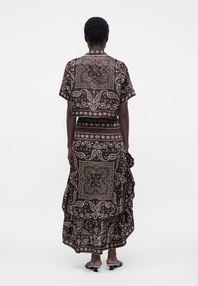 Personne portant un haut marron à manches courtes à motif paisley et une jupe midi à volants assortie, debout de dos sur fond blanc.
