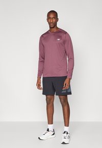 Bordeaux langtærmet atletisk skjorte med rund hals, parret med sorte shorts med blå stribe detaljer. Modellen bærer hvide sneakers.