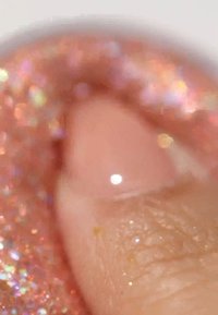 Close-up van een vingertop met een lichtroze glanzende nagellak met iriserende glitters in oranje en paarse tinten, die textuur benadrukken.