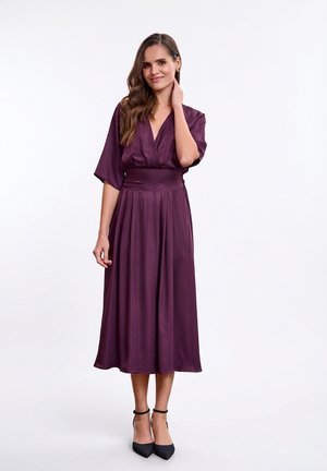 Fioletowa sukienka z satyny z głębokim dekoltem w kształcie litery V, krótkimi rękawami kimono oraz dopasowaną talią. Rozkloszowana spódnica o długości do kolan. Do tego pasujące czarne szpilki.
