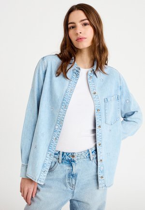 Młoda kobieta z długimi brązowymi włosami, ubrana w jasnoniebieską haftowaną kurtkę dżinsową na białej koszuli i dżinsy z wysokim stanem.