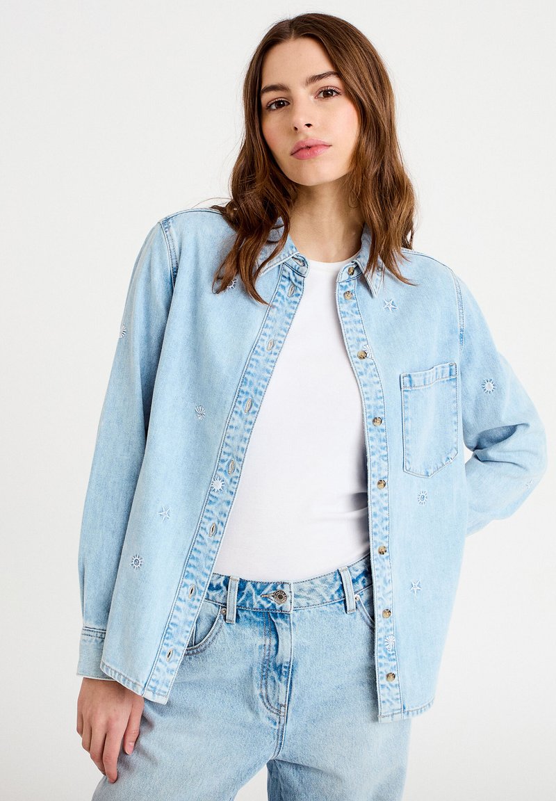 Giovane donna con capelli castani lunghi che indossa una giacca di jeans ricamata azzurro chiaro sopra una camicia bianca e jeans a vita alta.