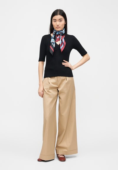 Femme debout, main sur la hanche, portant un haut noir à manches trois-quarts, un pantalon large beige, un foulard à motifs autour du cou et des chaussures marron.