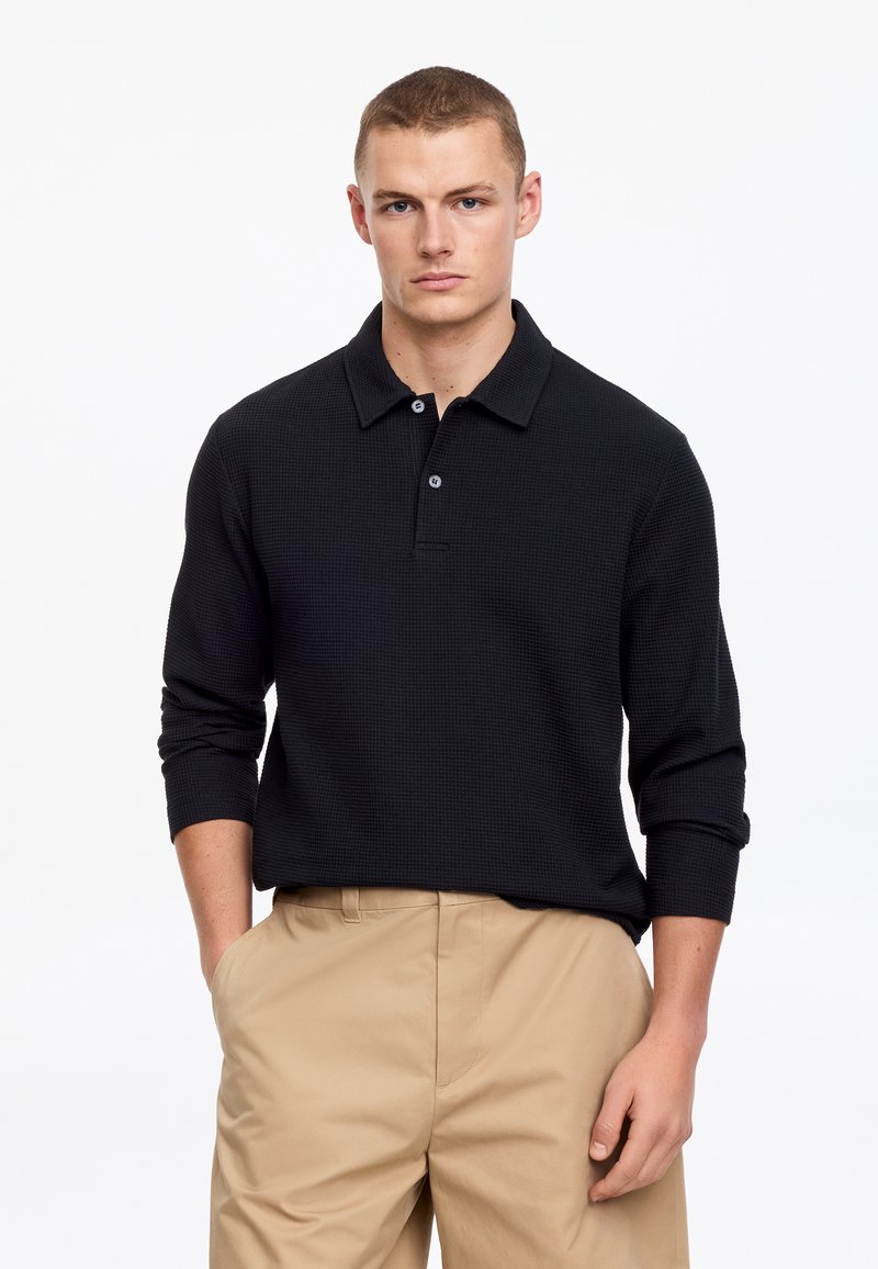 Joven con camisa polo negra de manga larga y pantalones beige, de pie con una mano en el bolsillo contra un fondo blanco.