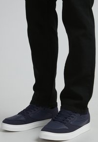 Navy blue sneakers with textured accents and lace closures, paired with black slim-fit pants. White rubber soles provide contrast.
