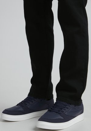 Navy blue sneakers with textured accents and lace closures, paired with black slim-fit pants. White rubber soles provide contrast.