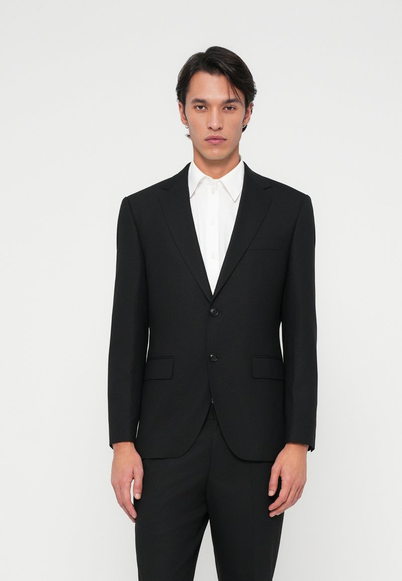 Veste de costume noire ajustée, à un seul bouton, avec des revers crantés et deux poches avant, portée sur une chemise blanche à col.