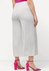 Pantalon blanc à jambes larges en tissu léger, présentant une texture lisse et une silhouette décontractée. Associé à des sandales à talons hauts roses.