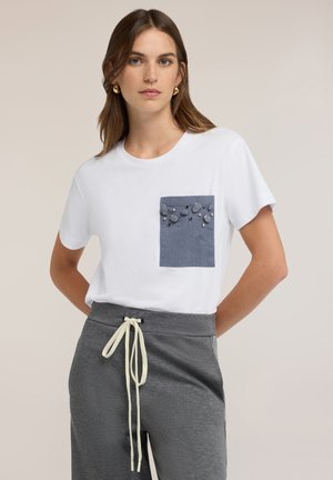 Frau trägt ein weißes T-Shirt mit blau verziertem Tasche und grauen Hose mit Tunnelzug, steht mit den Händen hinter dem Rücken.
