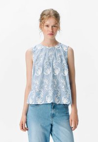 Top sin mangas de color azul claro con bordado floral blanco, escote redondo y dobladillo con forma de concha, combinado con jeans de denim claro.