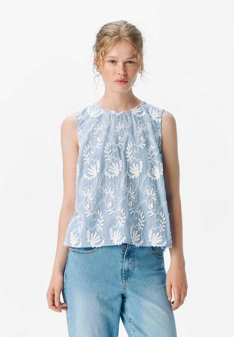 Top sin mangas de color azul claro con bordado floral blanco, escote redondo y dobladillo con forma de concha, combinado con jeans de denim claro.