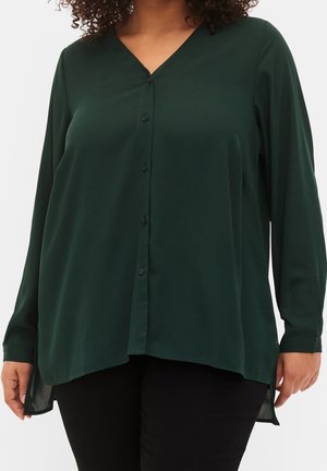 Vrouw draagt een donkergroene, langarmige blouse met knopen en V-hals, over een zwarte broek, met een ontspannen, losse pasvorm.