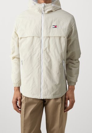 Mężczyzna w beżowej bluzie z kapturem na suwak, z elastycznymi mankietami i kieszeniami na zamek po bokach, w połączeniu z jasnobrązowymi spodniami.