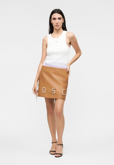 Γυναίκα με μακριά σκούρα μαλλιά, φορώντας λευκή αμάνικη μπλούζα, μίνι φούστα από ταμπά δέρμα με γράμματα από σκοινί και μαύρα πέδιλα με λουράκια, που στέκεται σε λευκό φόντο.