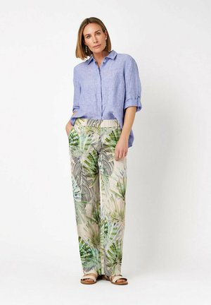 Frau steht mit Händen in den Taschen, trägt ein hellblaues Leinenhemd und weit geschnittene Hose mit grünem tropischem Blattmuster und Sandalen.