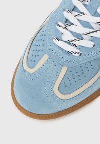 Μπλε σουέτ αθλητικό παπούτσι με μπεζ λεπτομέρειες, διάτρητη υφή, λευκά κορδόνια και εξωτερική σόλα από καουτσούκ χρώματος καραμέλας. Διαθέτει κυρτό σχεδιασμό.