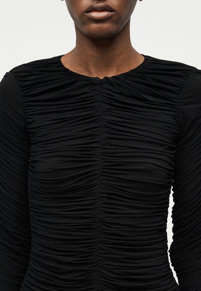 Detailní záběr osoby v černém tričku s dlouhým rukávem, které má na přední straně a rukávech shrnutý, nařasený materiál.