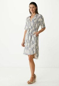 Vestido de manga corta con estampado en crema y líneas geométricas azules. Presenta una cintura ceñida con cinturón y bolsillos. Combinado con sandalias de tacón doradas.