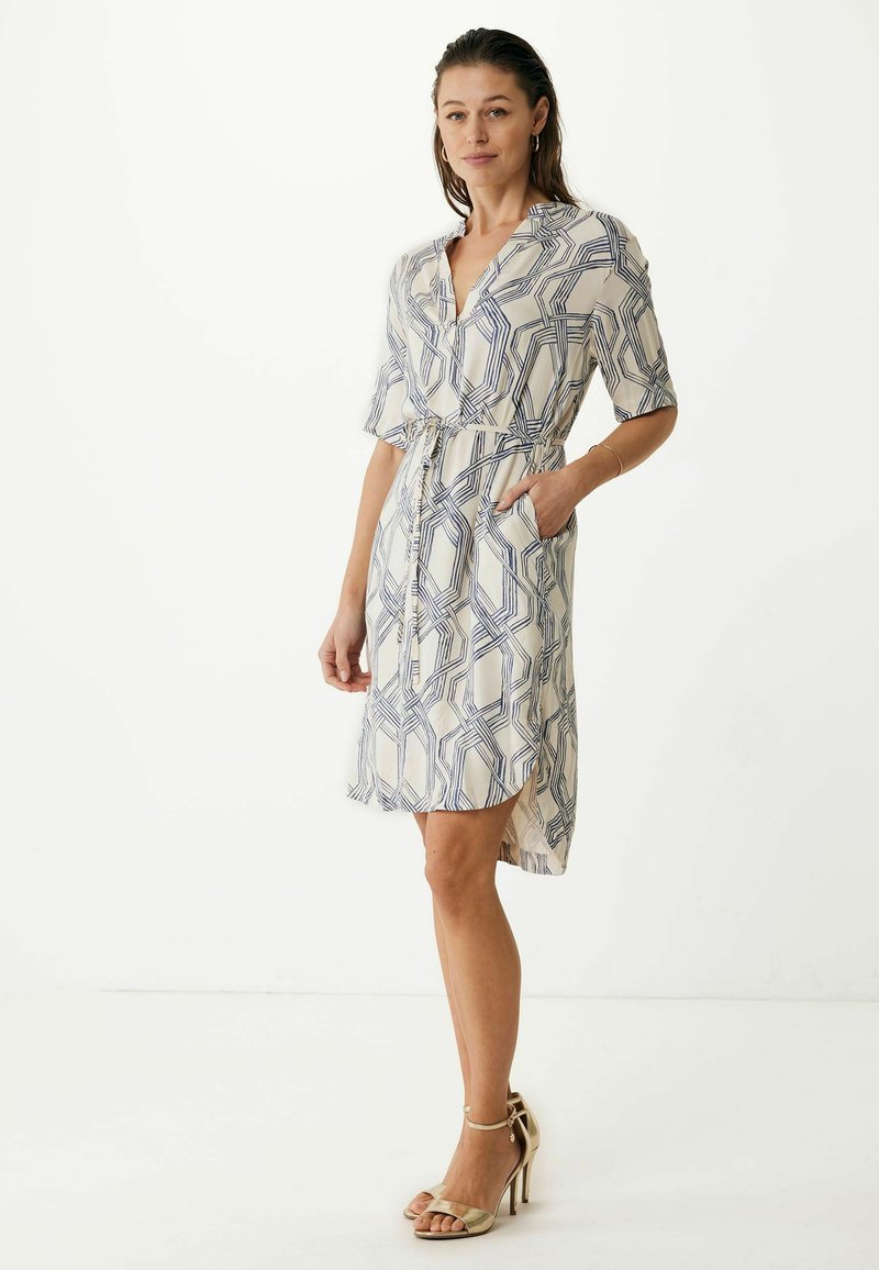 Vestido de manga corta con estampado en crema y líneas geométricas azules. Presenta una cintura ceñida con cinturón y bolsillos. Combinado con sandalias de tacón doradas.
