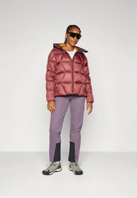 Bordowa pikowana kurtka puchowa z przeszyciami w kształcie diamentów, żółtym wnętrzem i zamkiem błyskawicznym z przodu. W zestawie z jasnoszarymi spodniami z czarnymi akcentami i szarymi butami.