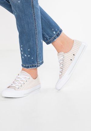 Sneaker beige chiaro con tomaia intrecciata testurizzata, suole in gomma bianca, sei occhielli in metallo e lacci bianchi, indossati con jeans blu strappati.