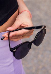 Černé sluneční brýle s velkými tmavými skly. Lesklé obruby mají tři tečky jako akcenty na spánku. Drží je ruka s prstenem.