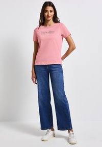 Helles rosa T-Shirt mit kurzen Ärmeln und einem grafischen Druck "Reflection", kombiniert mit weiten blauen Jeans und weißen Sneakers.